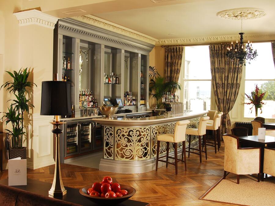 A bar area with a curved counter, bar stools, and a dining table with chairs. There are potted plants and a chandelier.
