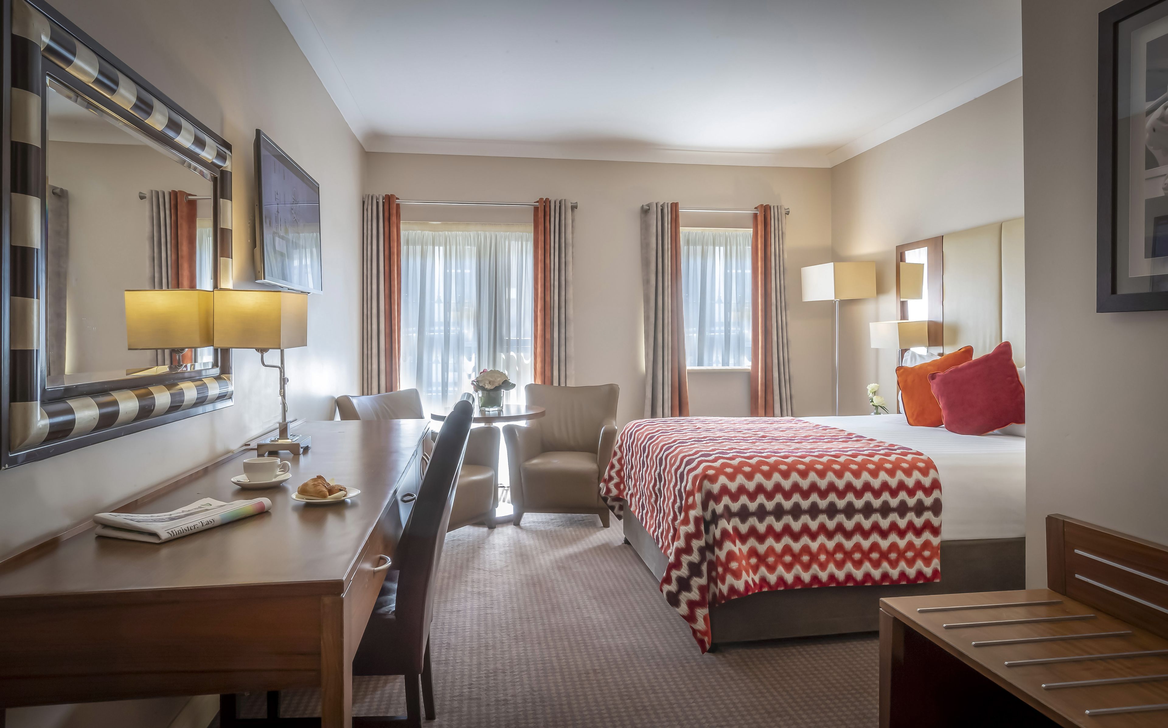 A hotel room with a bed, a desk, a chair, a lamp, and a mirror.