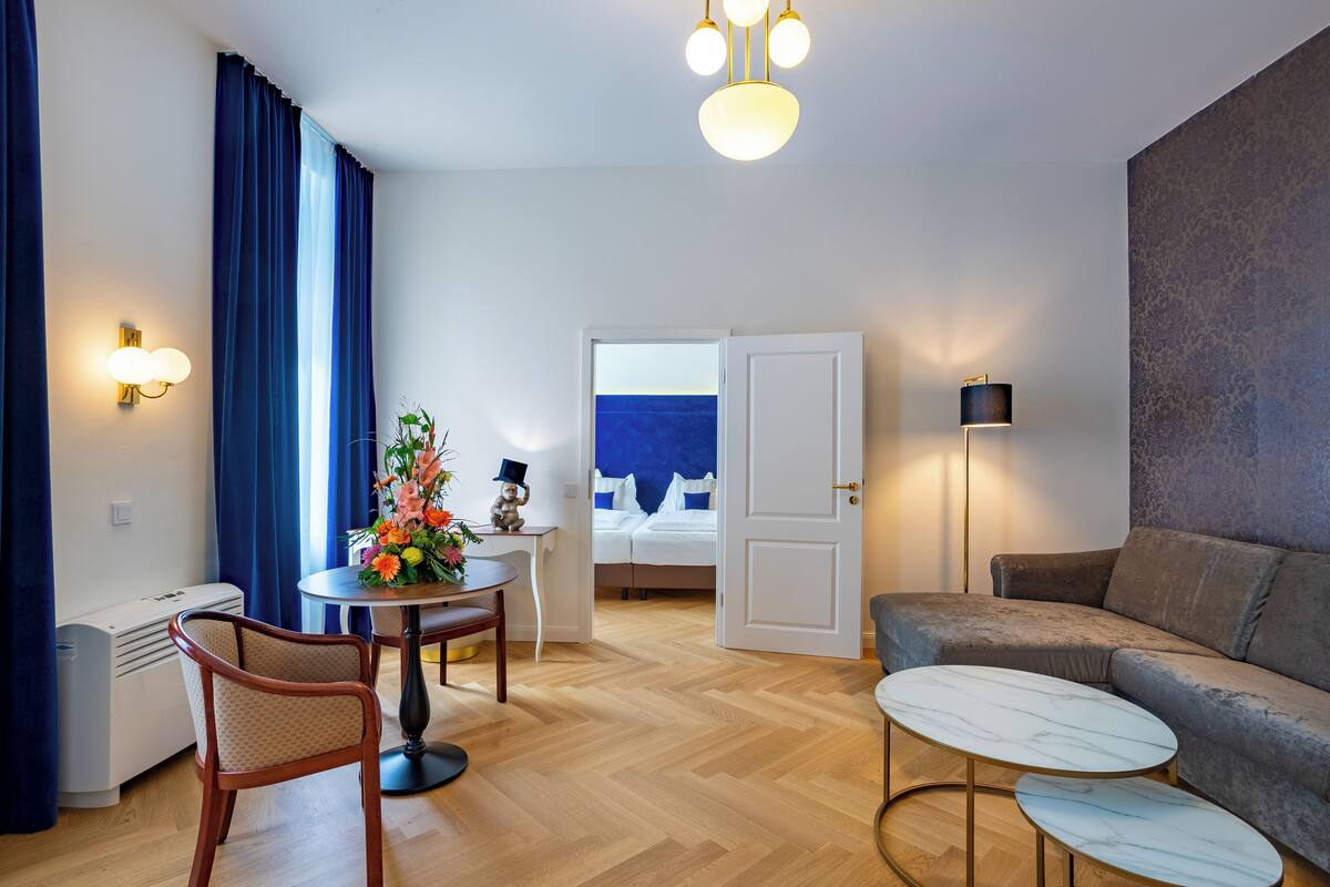 A modern hotel room with a wooden herringbone floor, a grey sofa, a round dining table with a flower centerpiece, a small side table with a lamp, and a view of a blue room through an open door.