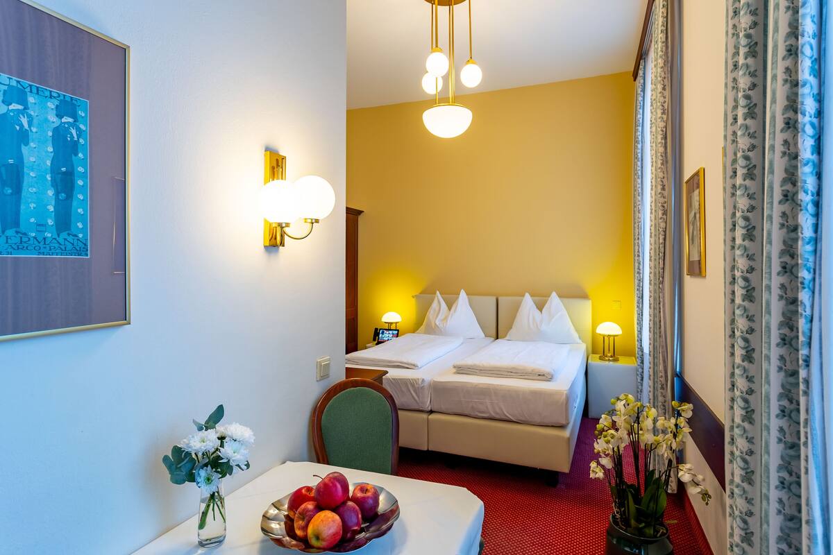 A hotel room with a bed, bedside tables, a desk with a bowl of fruit, a vase with flowers, and a poster on the wall.