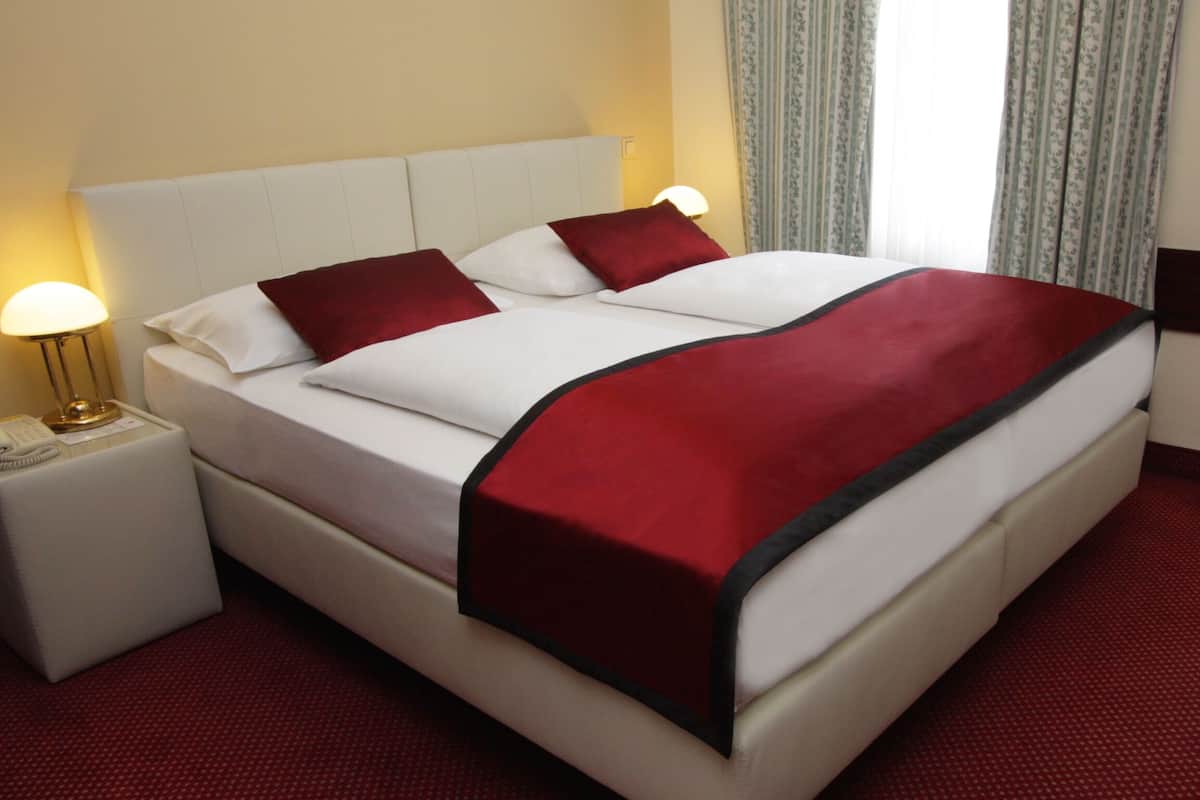 A hotel room with a bed, white headboard, red and white bedding, a red throw blanket, a bedside lamp, and a telephone on a white nightstand.