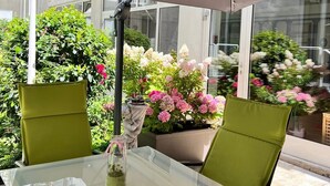 Un patio al aire libre con una mesa de vidrio y sillas verdes bajo un paraguas grande, rodeado de plantas y flores en macetas.