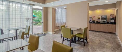 A modern dining area with tables and chairs, a kitchen area with a coffee machine, and a view through a glass door.