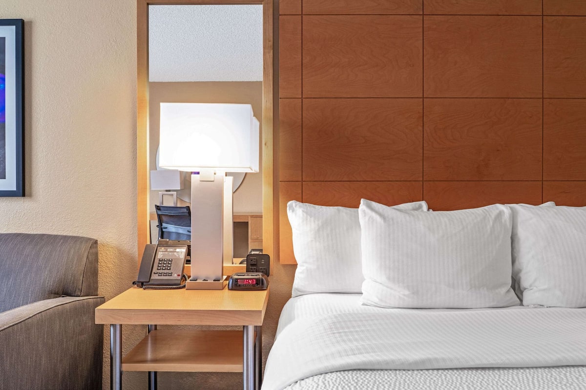 A hotel room with a bed, a bedside table with a lamp, a phone, and a clock.