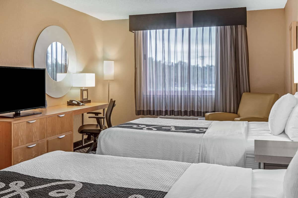 A hotel room with two beds, a desk, a television, and a large window with curtains.
