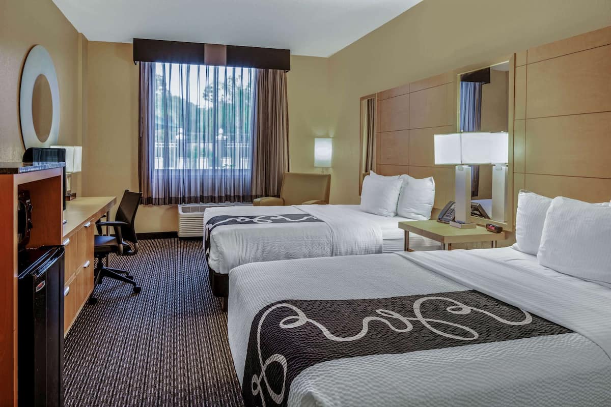 A hotel room with two beds, a desk, a chair, a TV, and a large window with curtains.