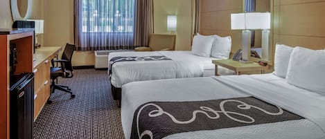 A hotel room with two beds, a desk, a chair, a TV, and a large window with curtains.