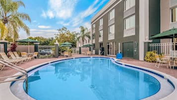 A swimming pool with lounge chairs and umbrellas, surrounded by a building with large windows and a paved walkway.
