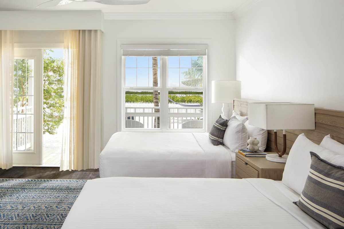 A hotel room with a large bed, a nightstand with a lamp, and a view of a white fence and greenery outside.
