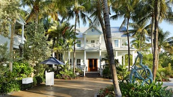 A two-story white building with a covered porch, surrounded by palm trees and tropical plants.
