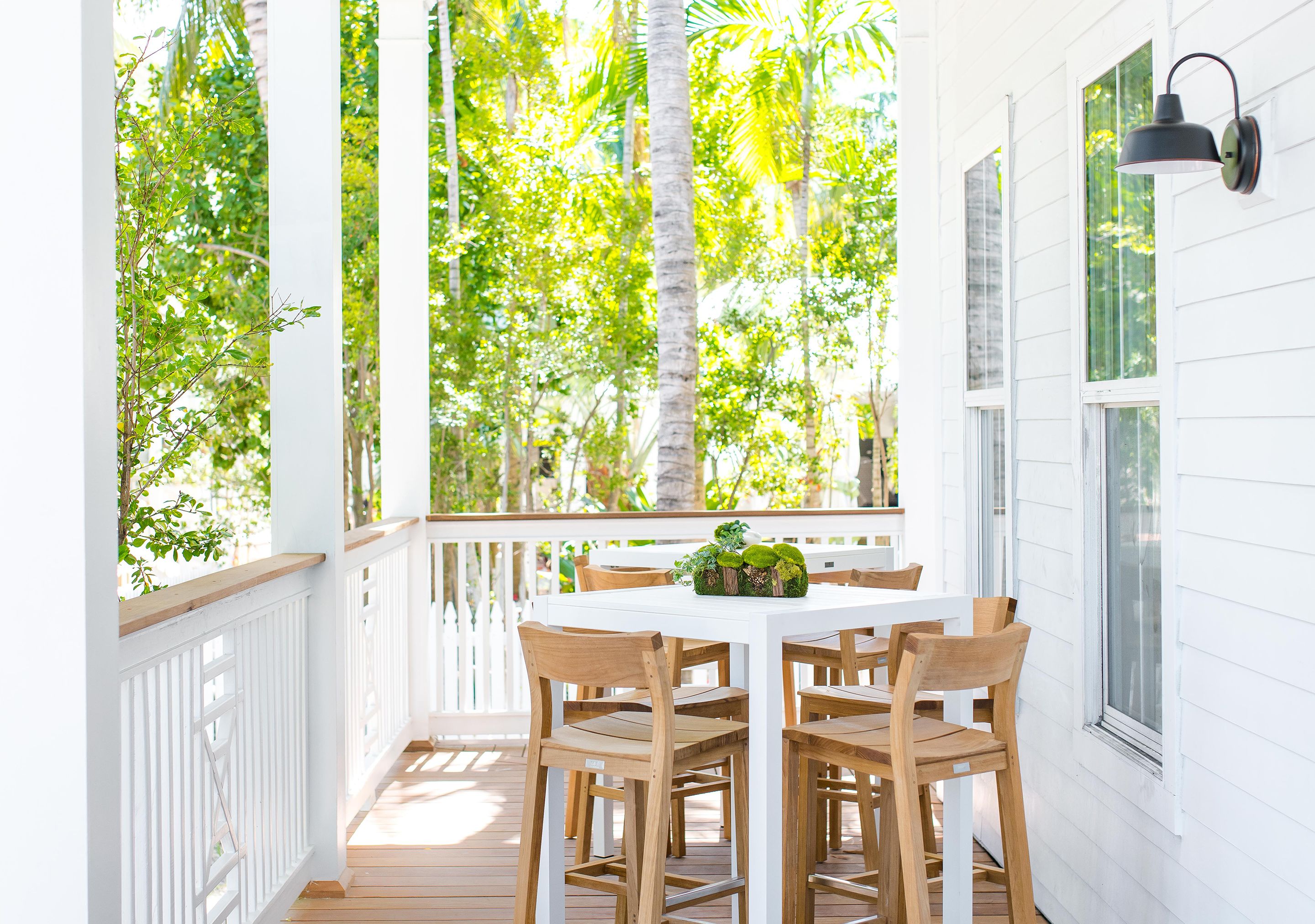 A white porch with a wooden table and chairs, surrounded by white railings and palm trees.