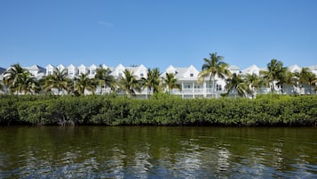 Una hilera de villas blancas frente a la playa, con palmeras, un cuerpo de agua al frente y un cielo azul despejado.