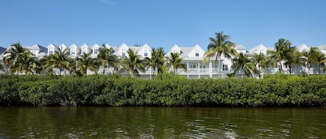 A row of white beachfront villas with palm trees, a water body in the foreground, and a clear blue sky.