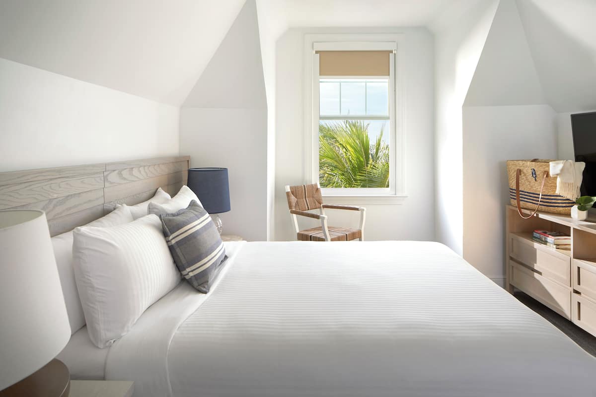 A bedroom with a large bed, a wooden headboard, a window with a view of palm trees, a chair, and a TV stand with a plant.
