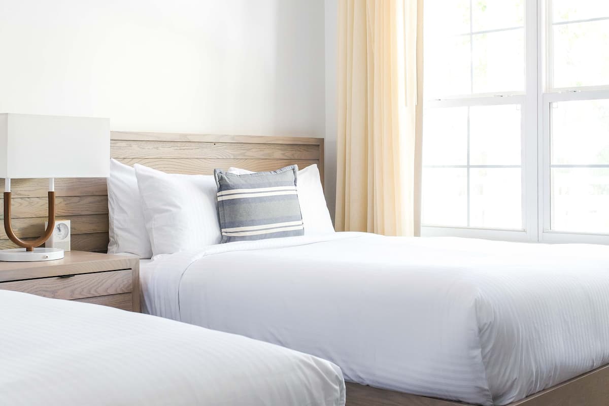 A hotel room with two beds, a bedside table with a lamp, and a window with curtains.