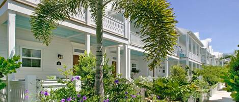 Hilera de casas blancas frente a la playa con balcones, rodeadas de exuberante vegetación y flores moradas.