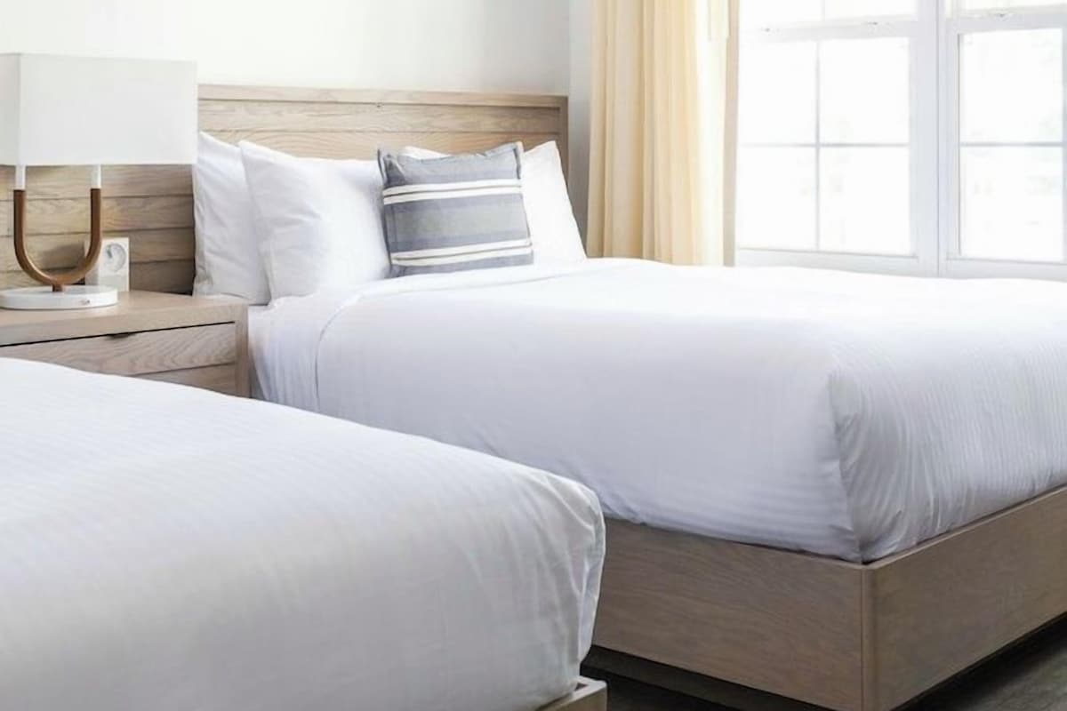 A hotel room with two beds, a bedside table with a lamp, and a window with curtains.