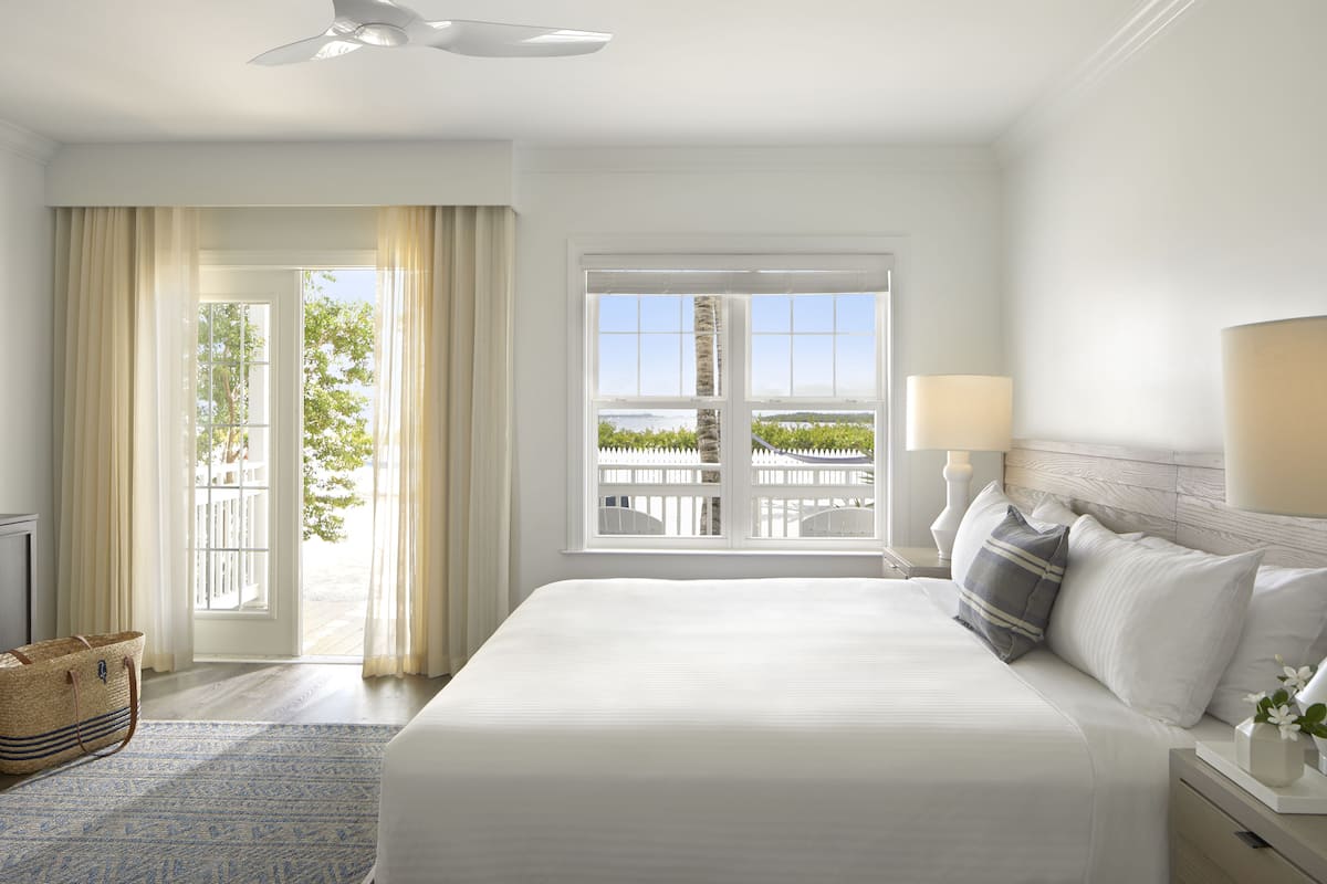 A hotel room with a large bed, a view of a white fence and trees, a ceiling fan, and a nightstand with a lamp.
