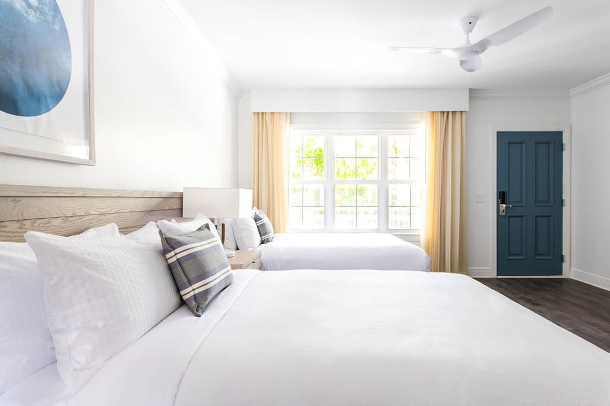 A hotel room with two beds, a ceiling fan, a window with curtains, and a blue door.