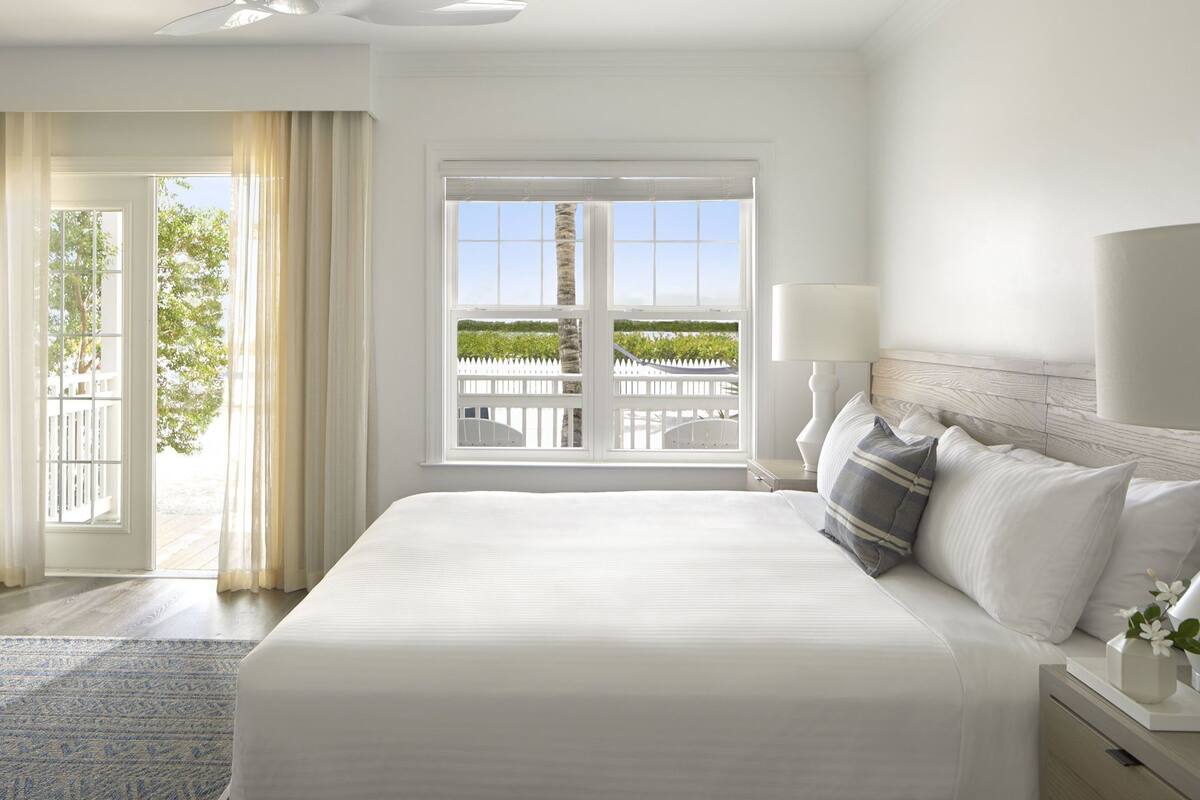 A hotel room with a large bed, a view of a white fence and greenery, and a balcony with a railing.
