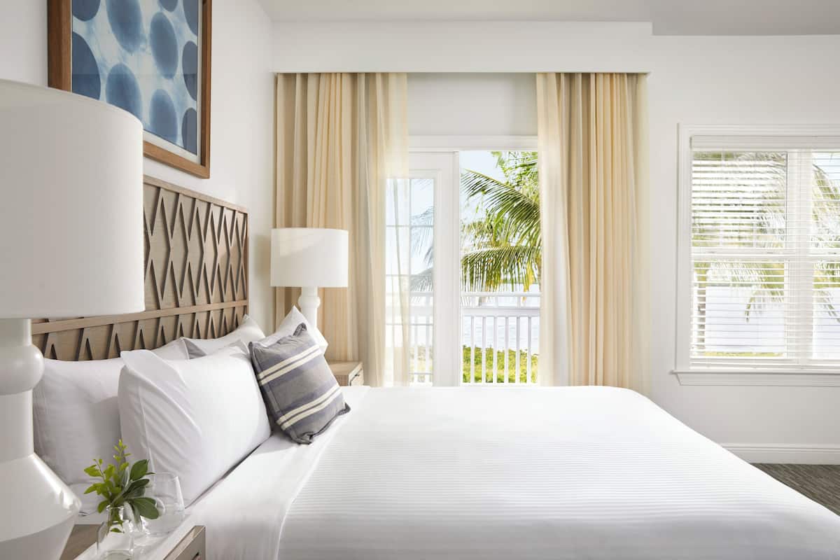 A hotel room with a large bed, a bedside table with a lamp, a view of the outdoors through a door, and a framed picture on the wall.
