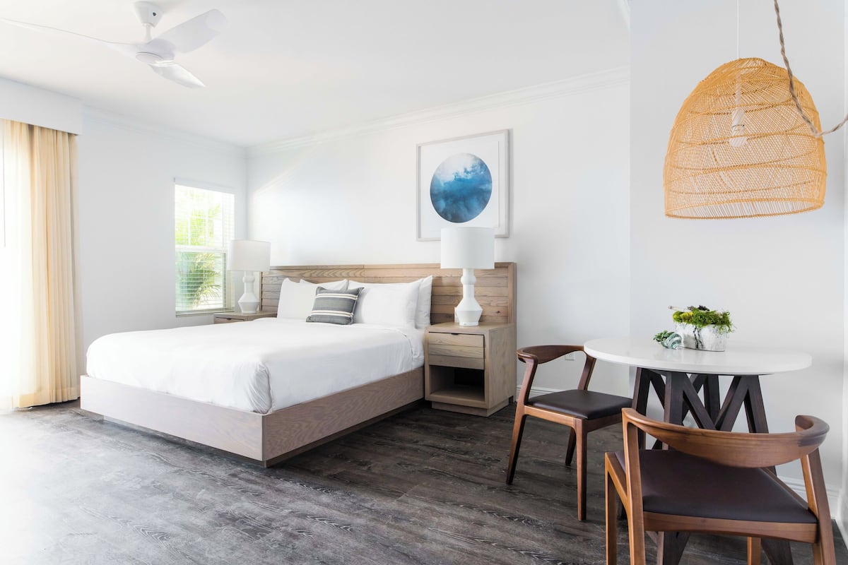 A modern hotel room with a large bed, bedside tables, a round dining table, and a ceiling fan.