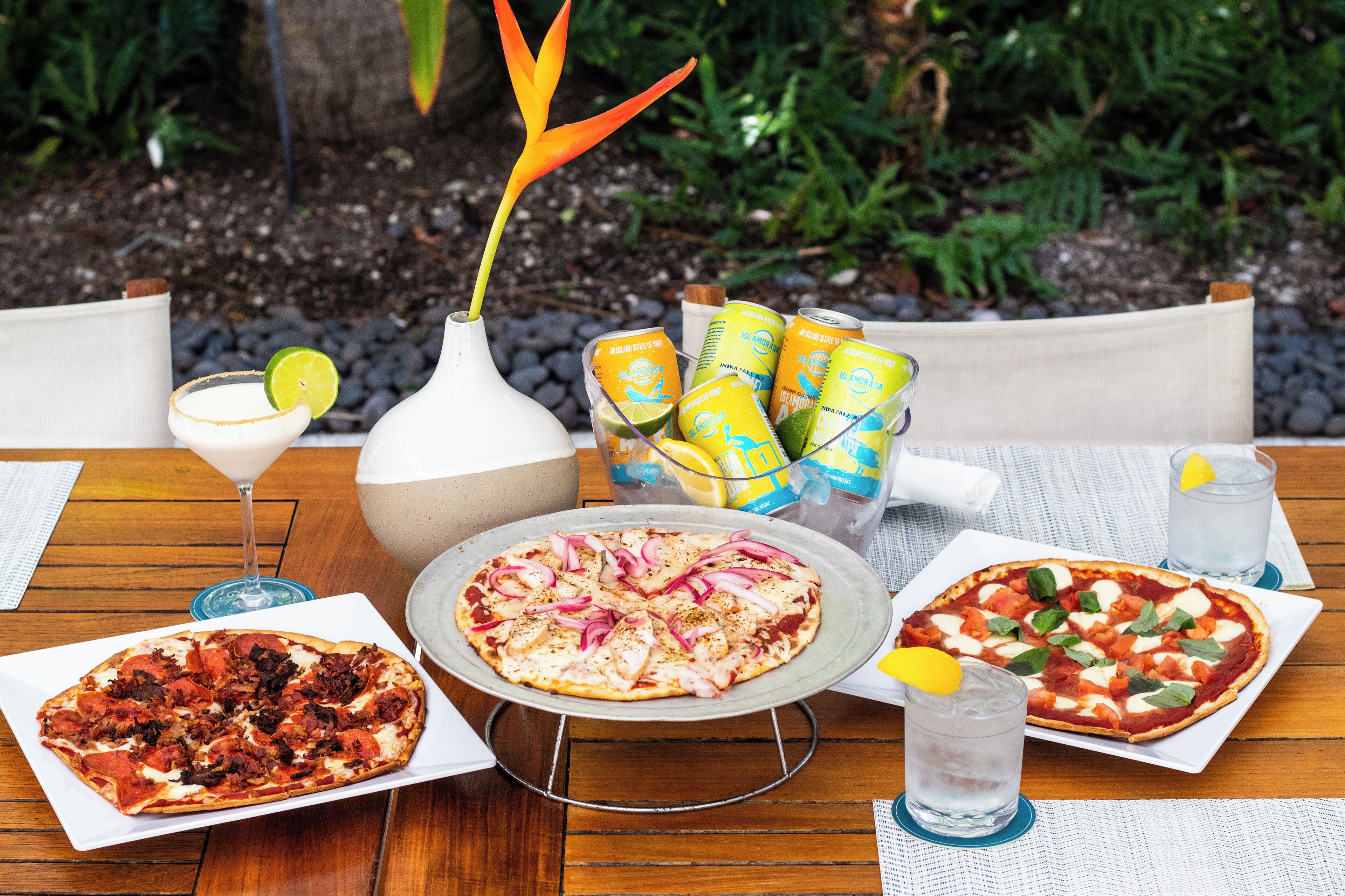 A table with three pizzas, a vase with a plant, and a beverage cooler with drinks.