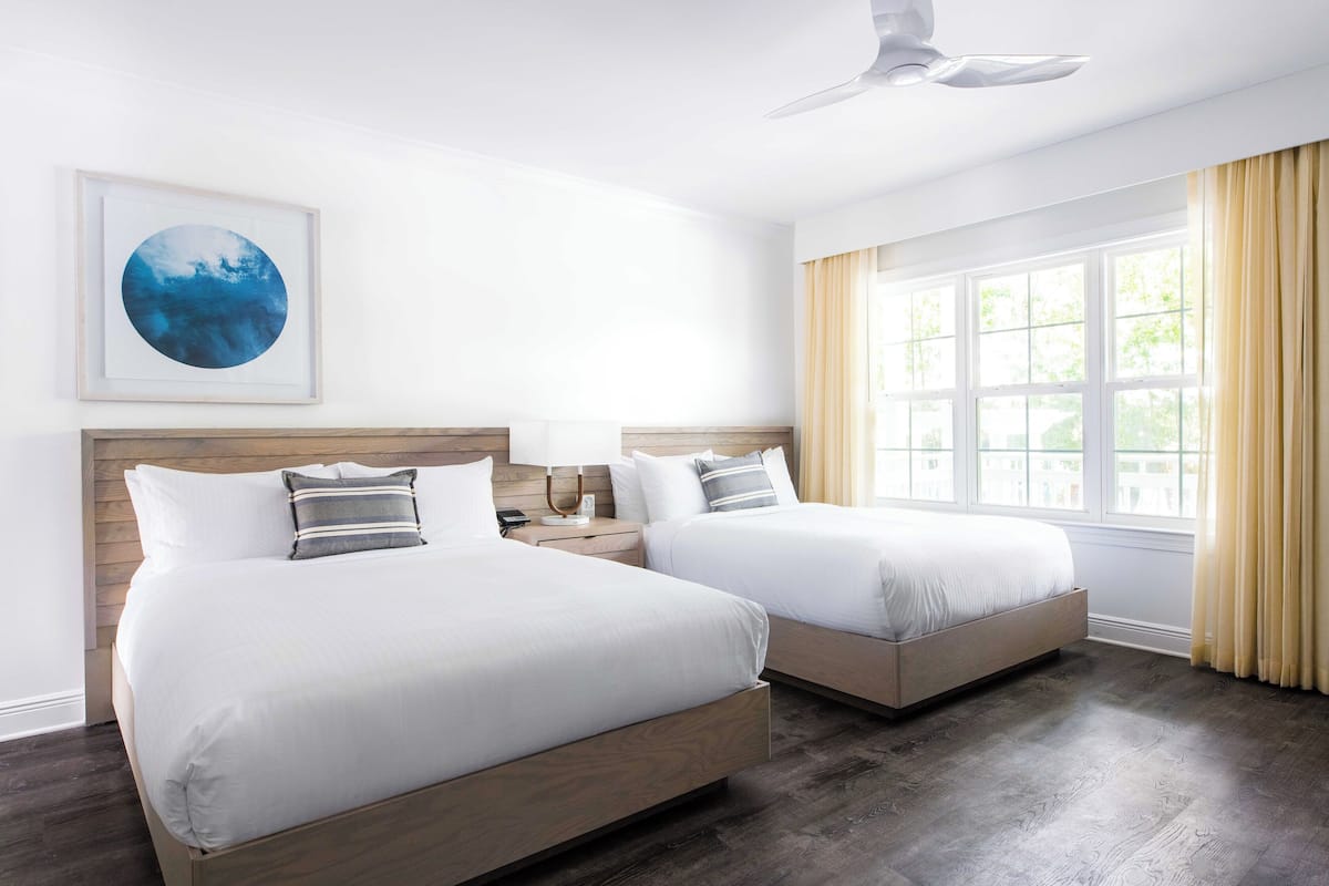 A hotel room with two beds, a ceiling fan, a large window with curtains, and a framed picture on the wall.