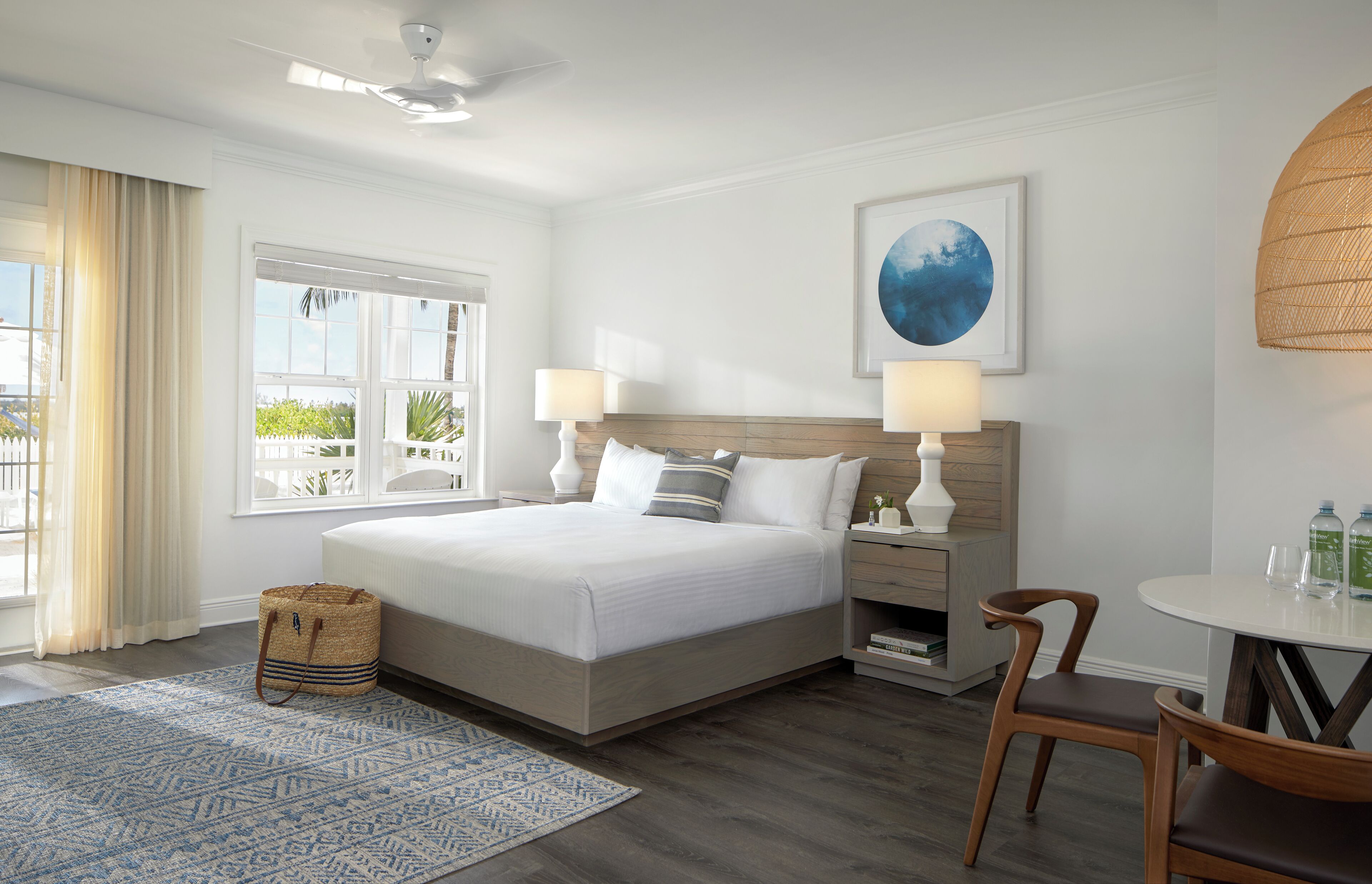 A hotel room with a large bed, two bedside tables with lamps, a small round table with chairs, a wicker basket, and a large window with curtains.