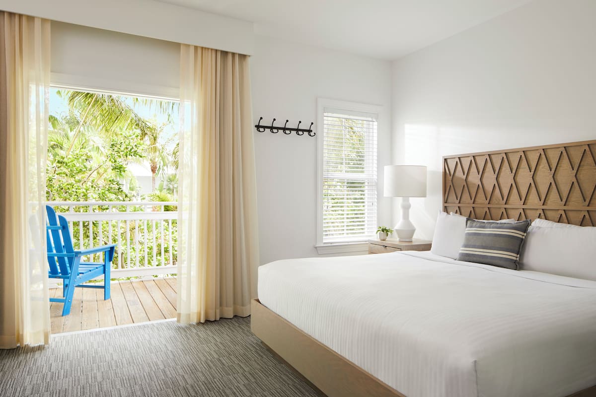A hotel room with a bed, a chair, a window with blinds, and a view of greenery outside.
