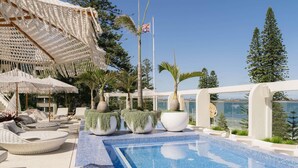 A rooftop infinity pool with lounge chairs and umbrellas, surrounded by potted plants and overlooking a body of water. - The Brighton Hotel Sydney - MGallery Collection (Brighton-le-Sands)