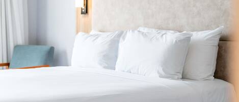 Una cama bien tendida con ropa blanca, un cabecero de tono claro y una mesita de noche con una lámpara.