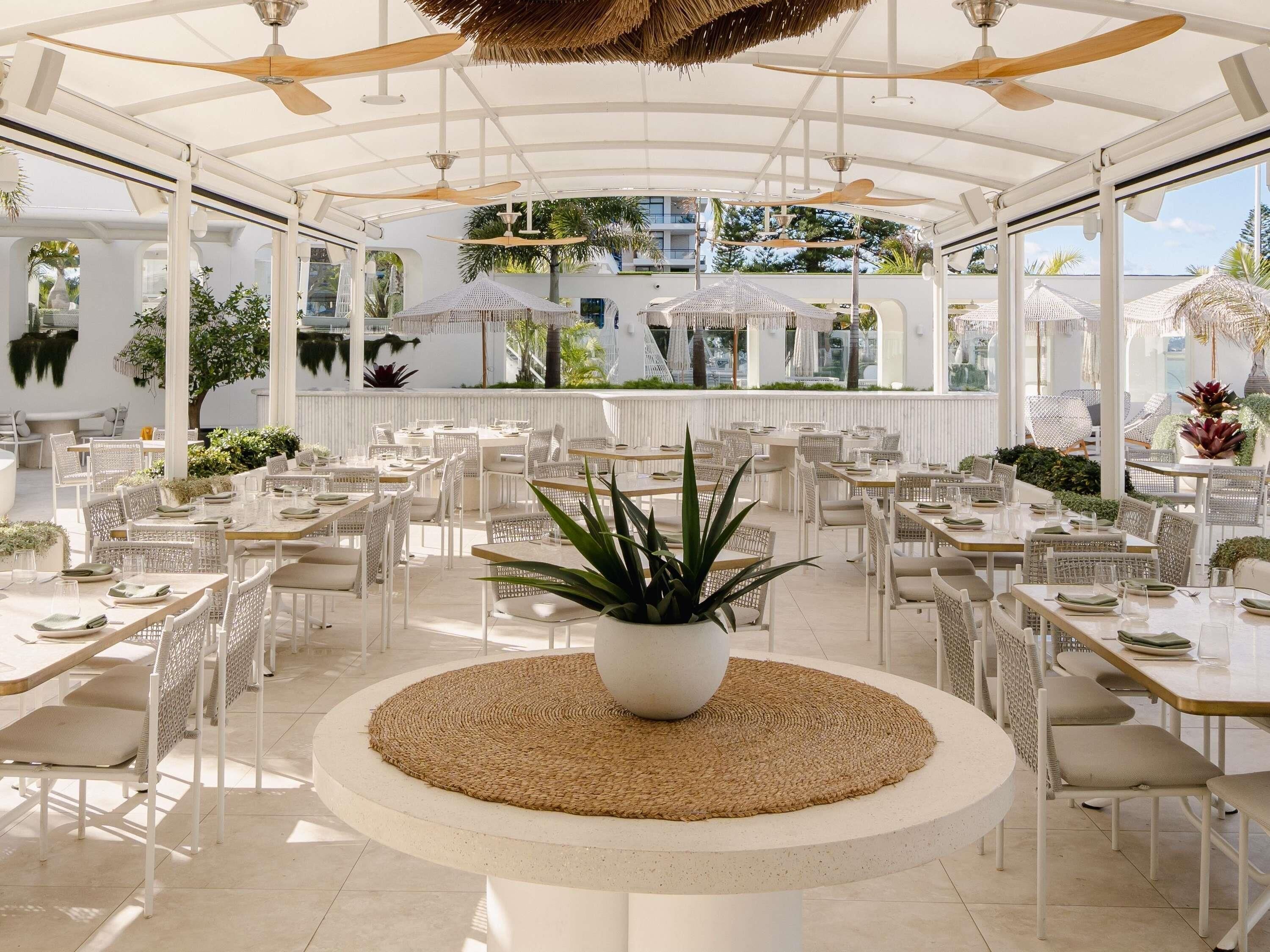 An open-air dining area with white furniture, ceiling fans, and potted plants.