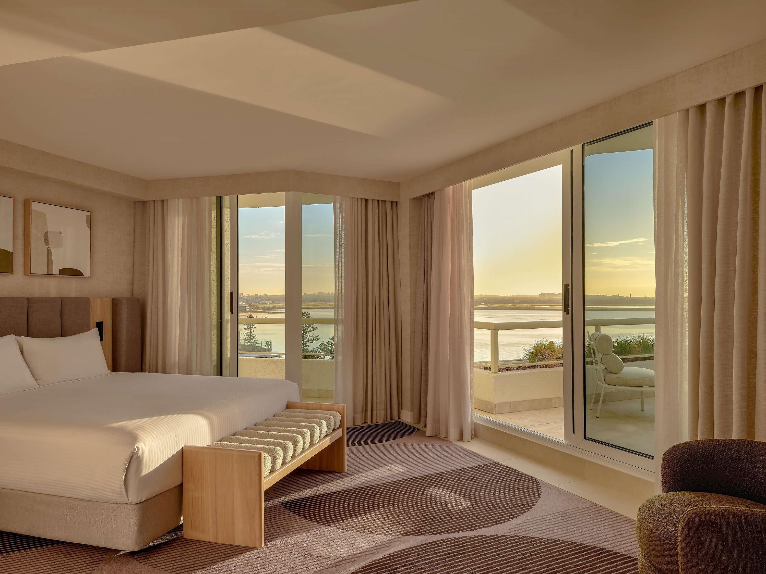 A hotel room with a large bed, a bench, a view of a body of water, and a balcony with curtains.