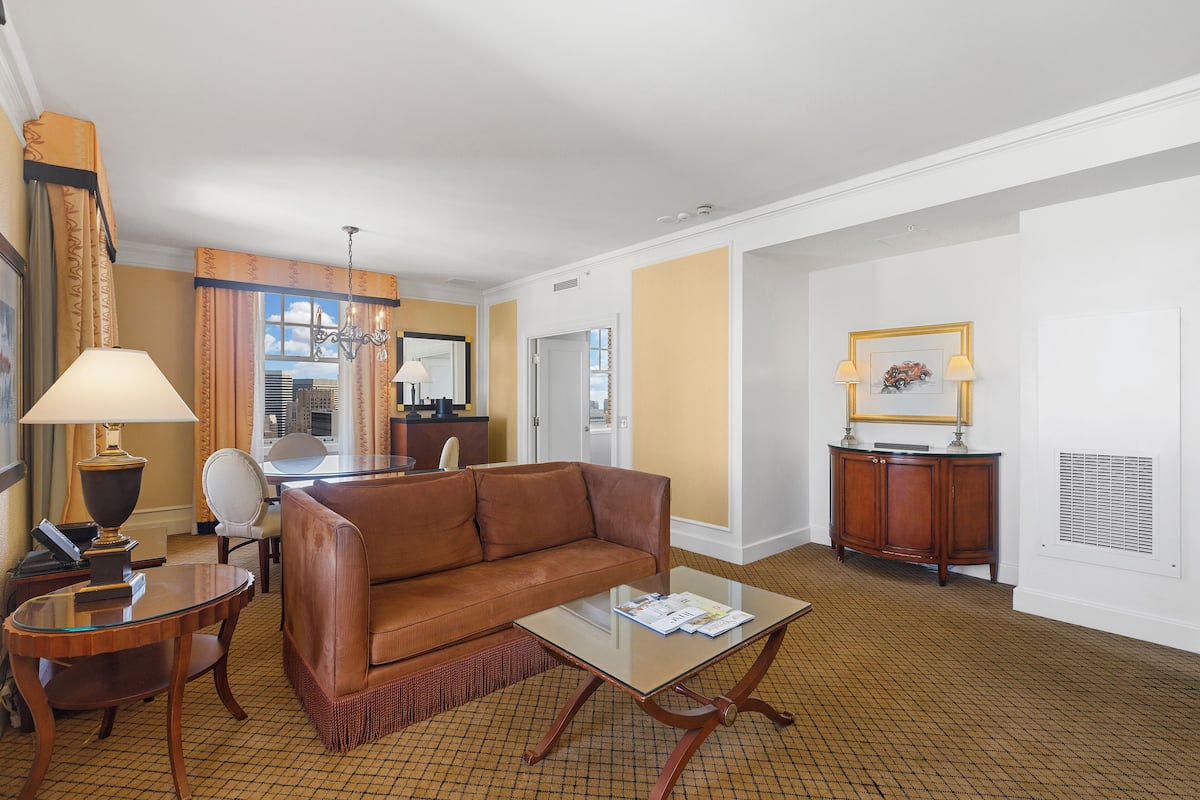 A hotel room with a brown leather sofa, a glass coffee table, a wooden side table, a lamp, a desk with a chair, a mirror, and a framed picture on the wall.