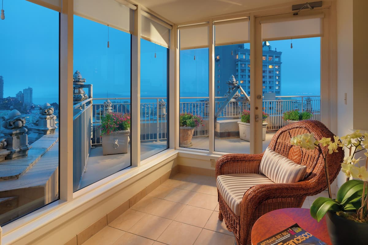 A balcony with a wicker chair and potted plants, large windows offering a city view, and a tiled floor.