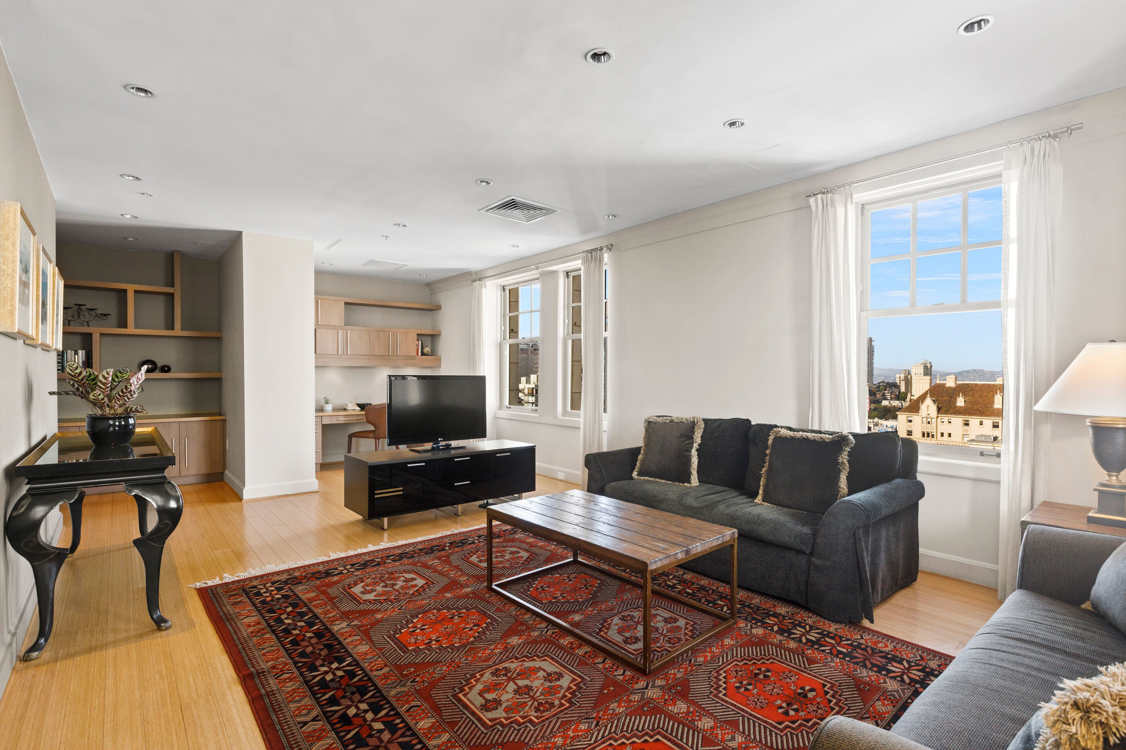 Una sala de estar moderna con un sofá, una mesita de centro y un televisor. Cuenta con un amplio ventanal con cortinas, una alfombra estampada y vistas a edificios.