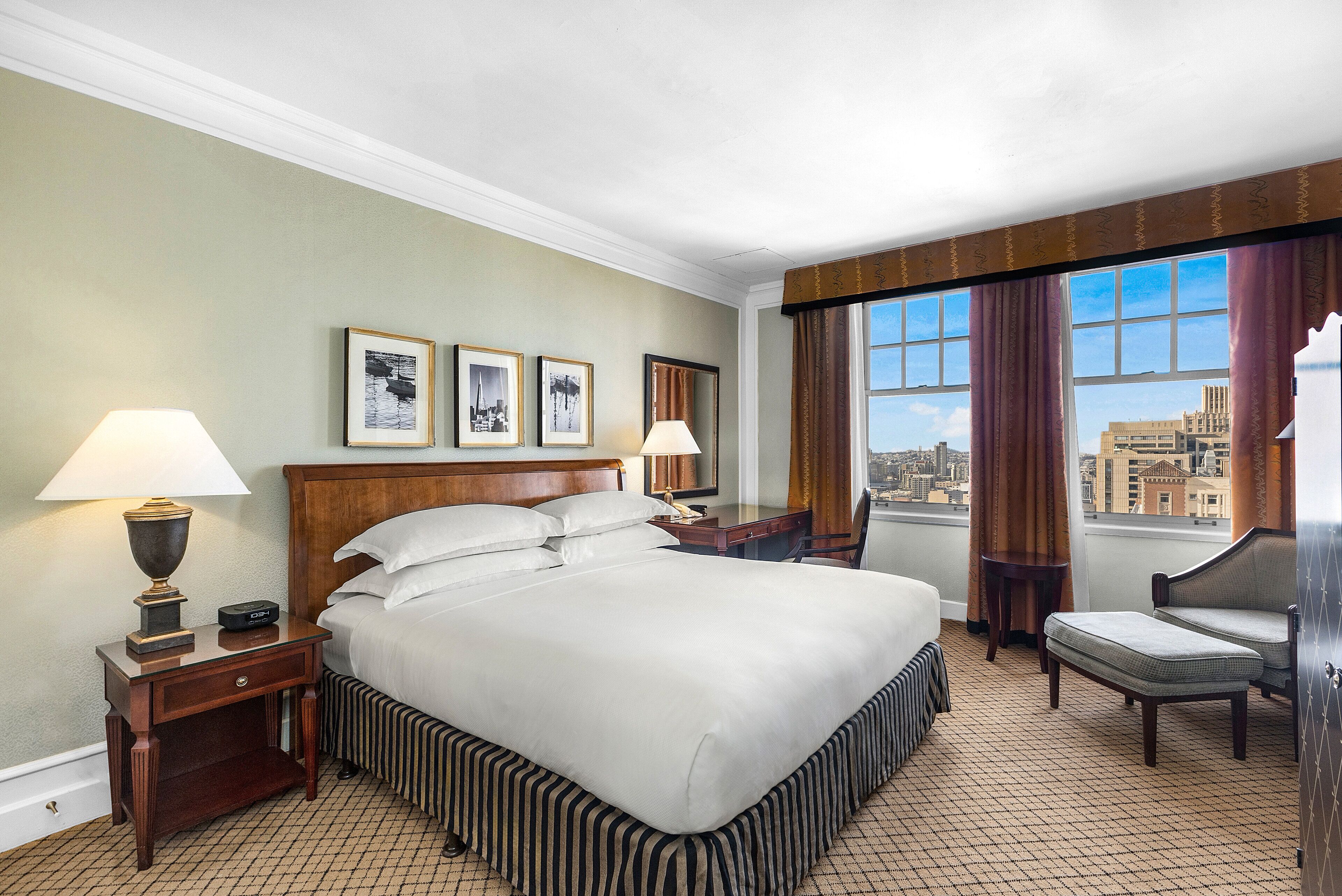 Ein Hotelzimmer mit einem großen Bett, zwei Nachttischen inklusive Lampen, einem Sessel und Blick auf die Stadt durch große Fenster.