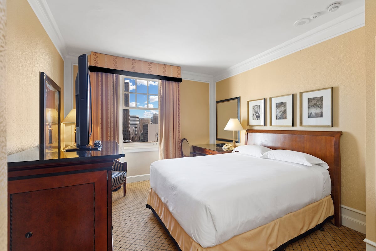 A hotel room with a large bed, a desk with a lamp, a chair, a mirror, and framed pictures on the wall.