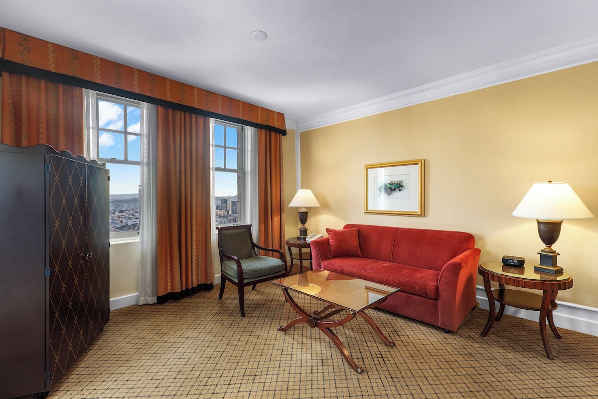 A hotel room with a red sofa, a wooden coffee table, a chair, a lamp, and a view of the city through the window.