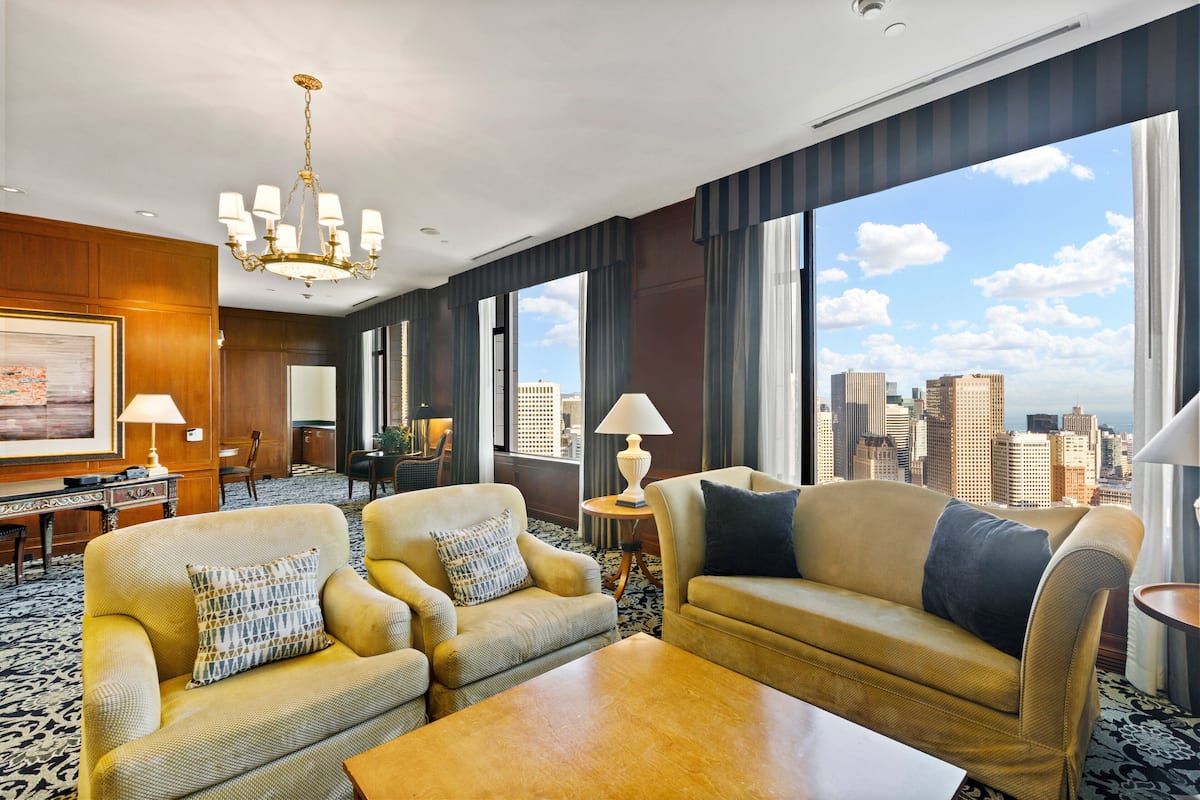 A hotel room with two sofas, a coffee table, a desk with a lamp, and large windows offering a city view.