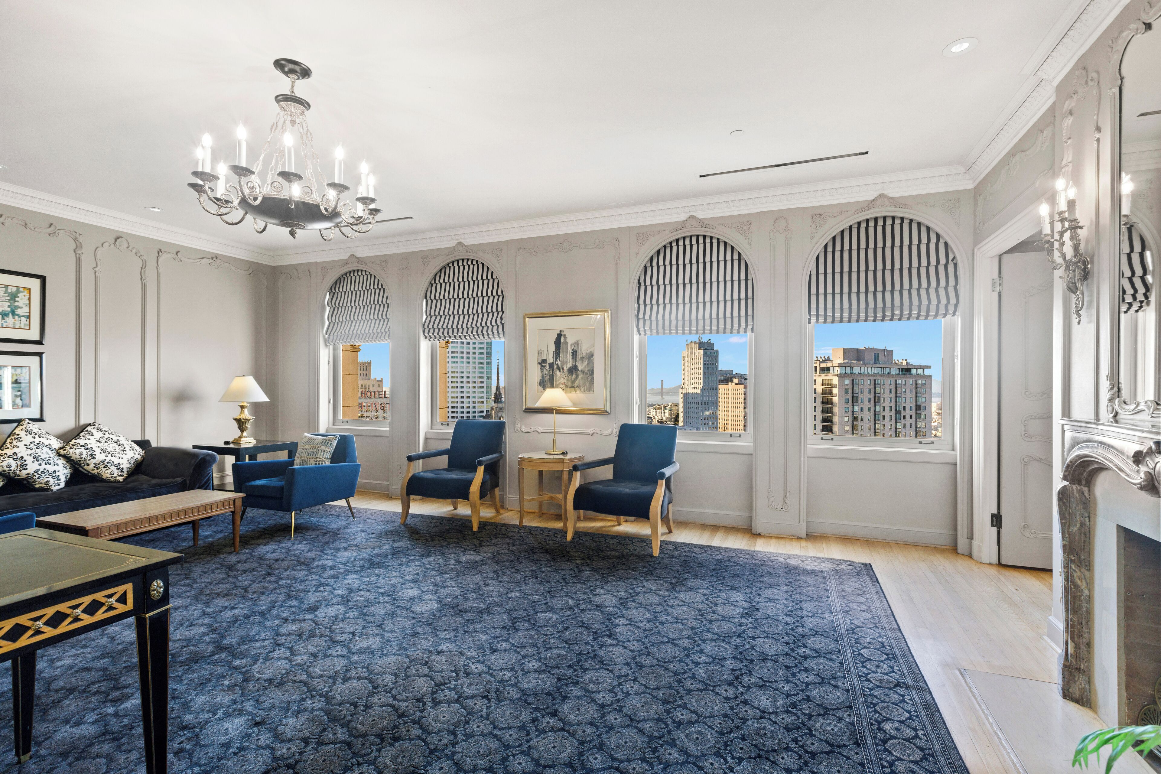 A spacious room with a patterned carpet, a chandelier, a fireplace, and a view of the city through large windows.