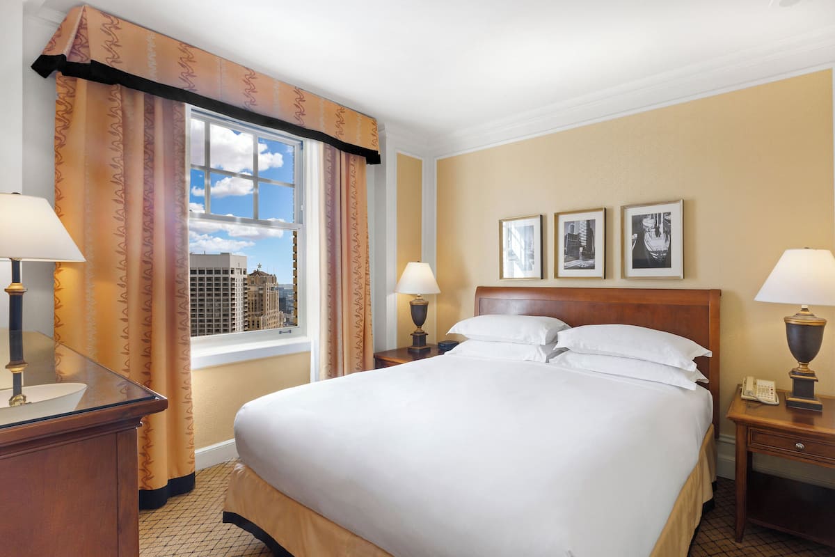 A hotel room with a large bed, two bedside tables with lamps, a window with curtains, and two framed pictures on the wall.