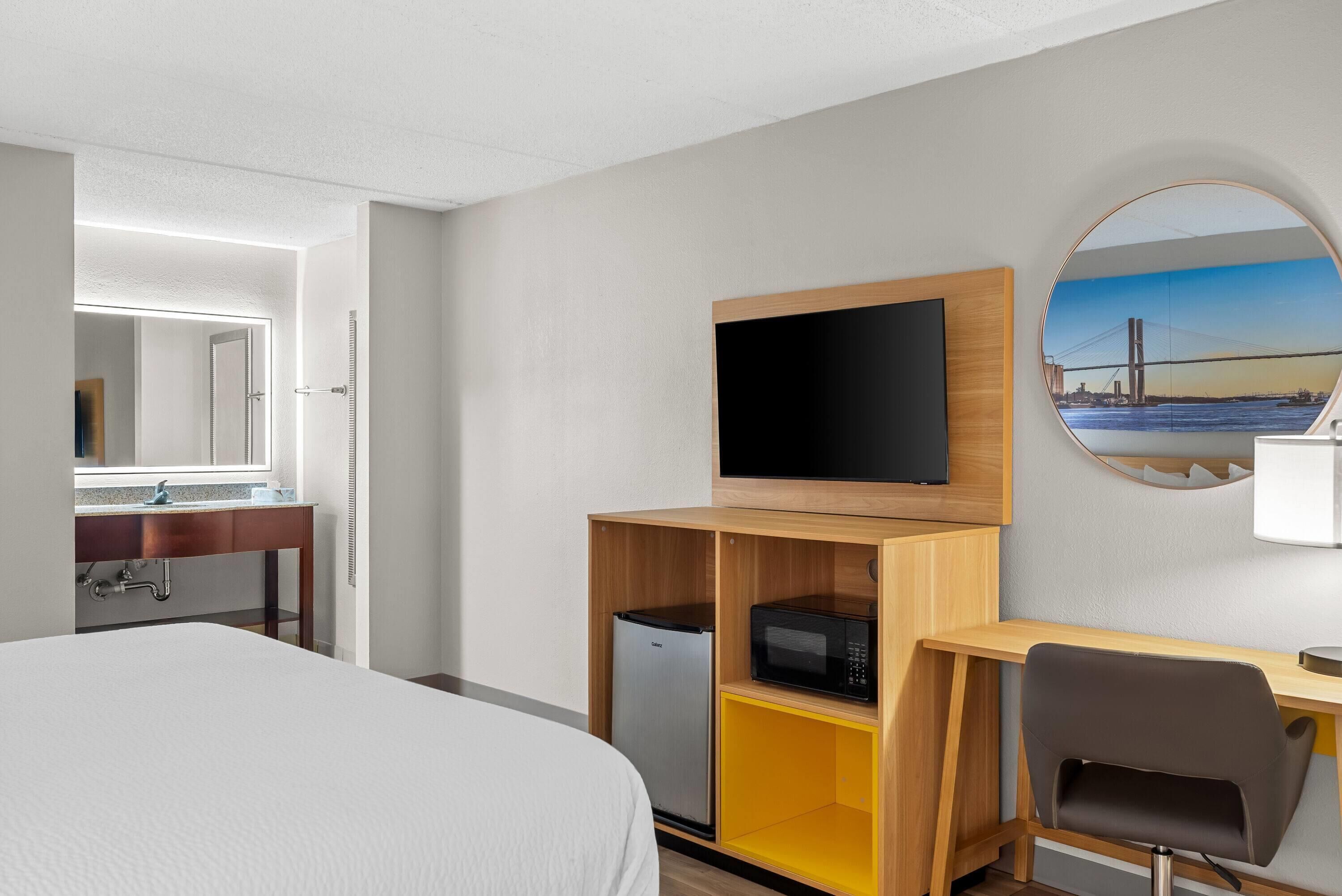 A hotel room with a bed, a desk with a computer, a microwave, and a TV on the wall.