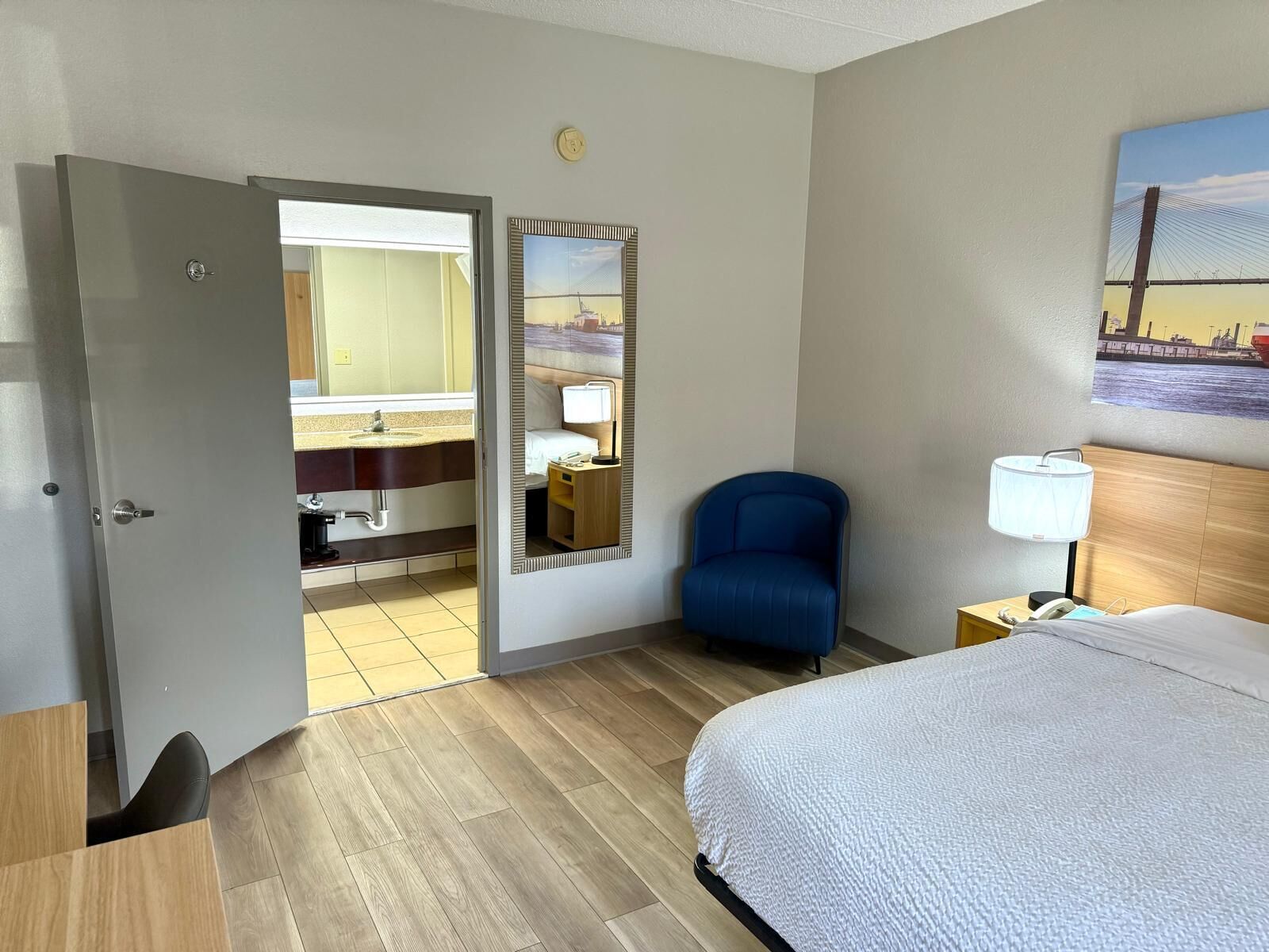 A hotel room with a bed, a desk, a chair, a nightstand, a lamp, a bathroom with a sink and mirror, and a painting on the wall.