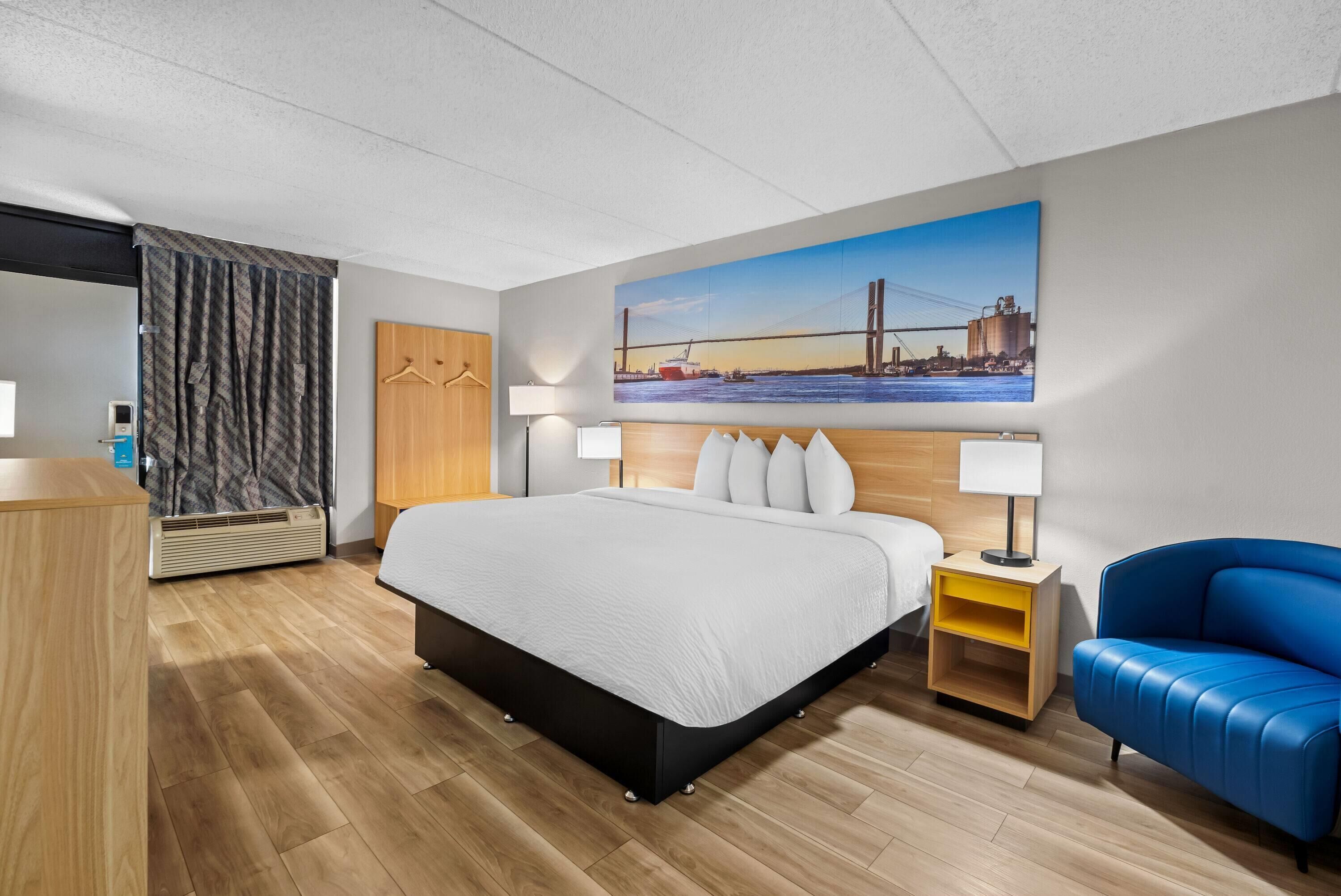 A modern hotel room with a large bed, a blue sofa, a wooden desk, and a painting of a bridge on the wall.