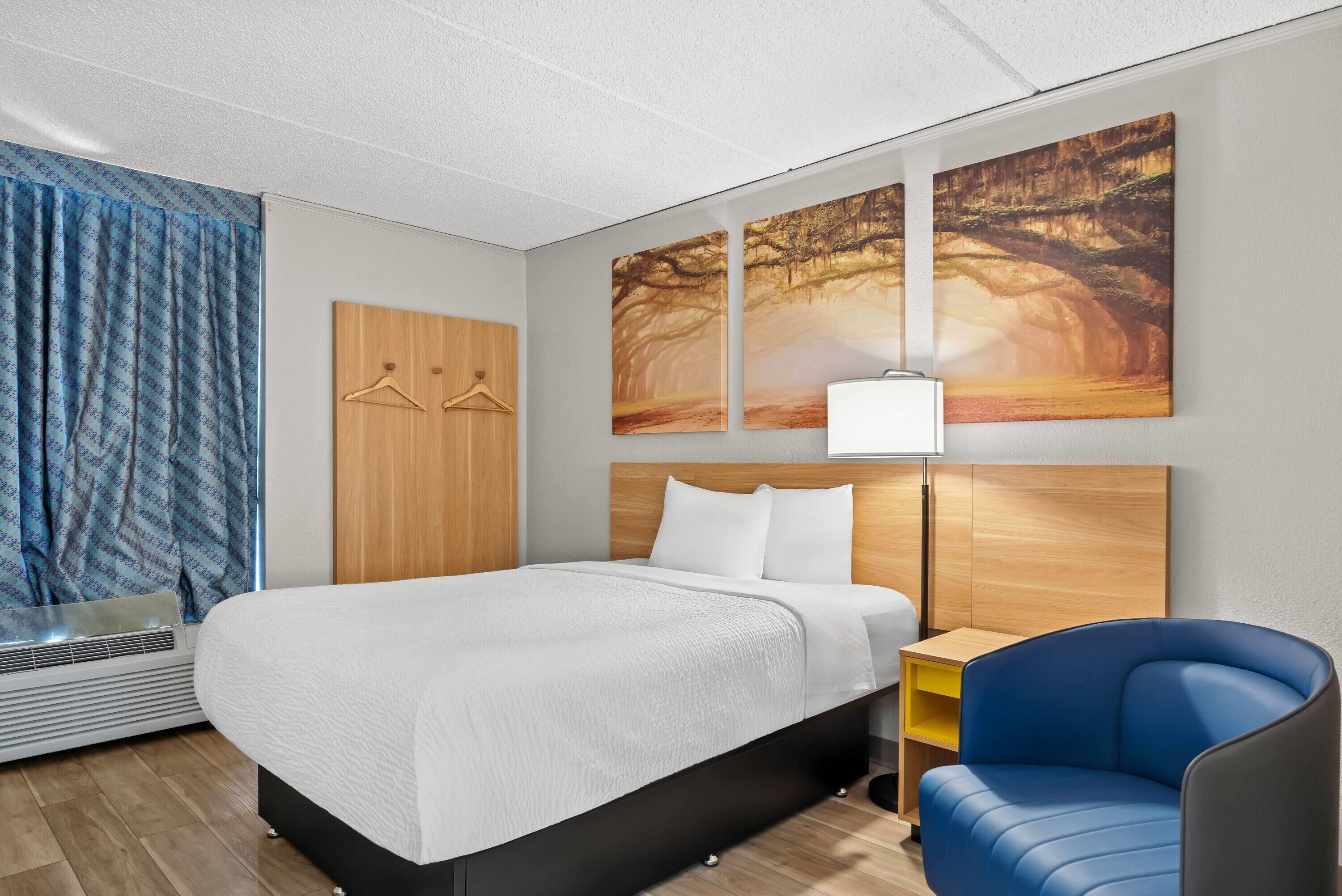 A modern hotel room with a large bed, a blue armchair, a wooden wardrobe, and three wall-mounted paintings.