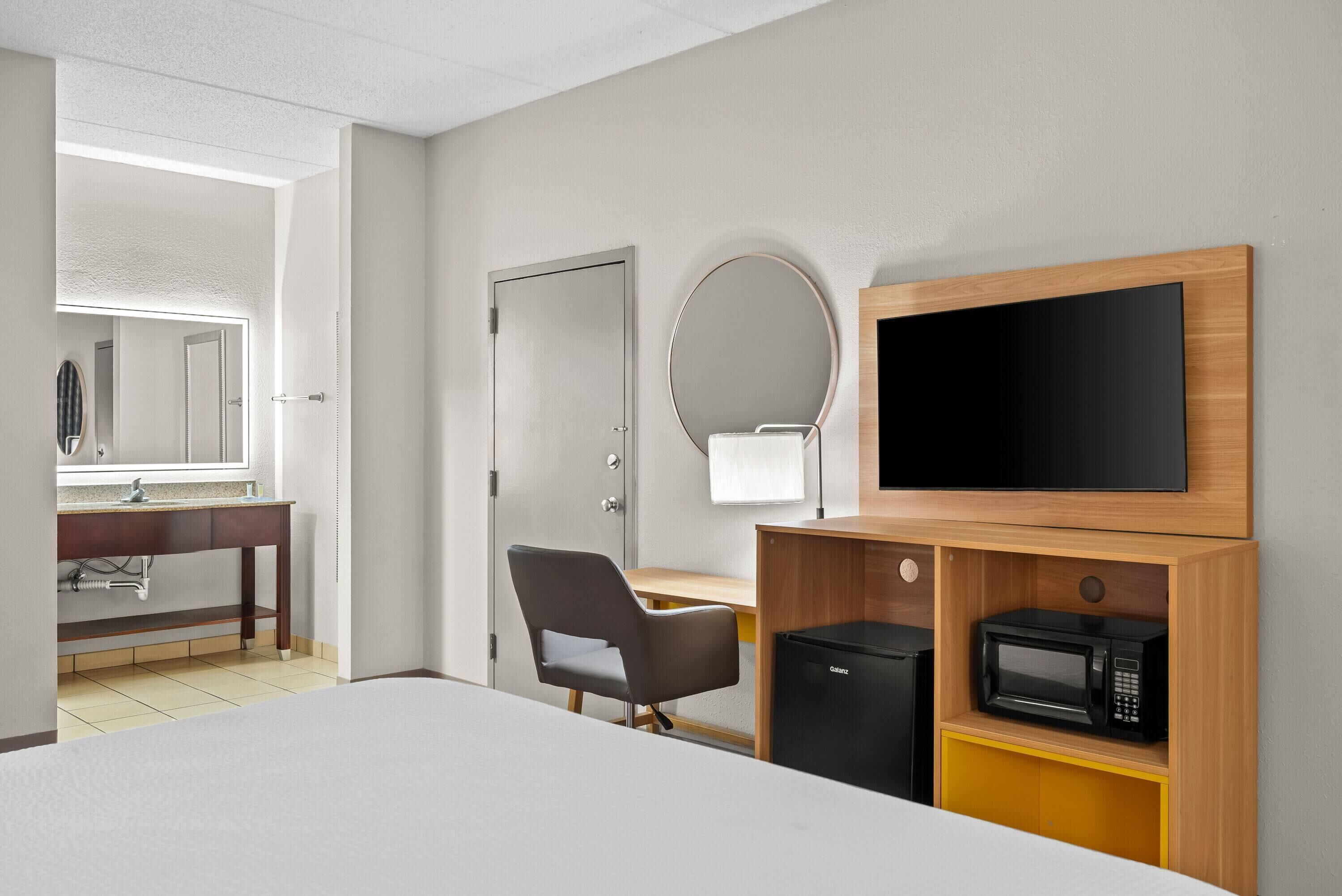A hotel room with a bed, a desk, a chair, a TV, a microwave, and a bathroom visible through an open door.