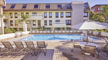 Zona de piscina de hotel con tumbonas, una mesa con sombrilla y vista al exterior del edificio.