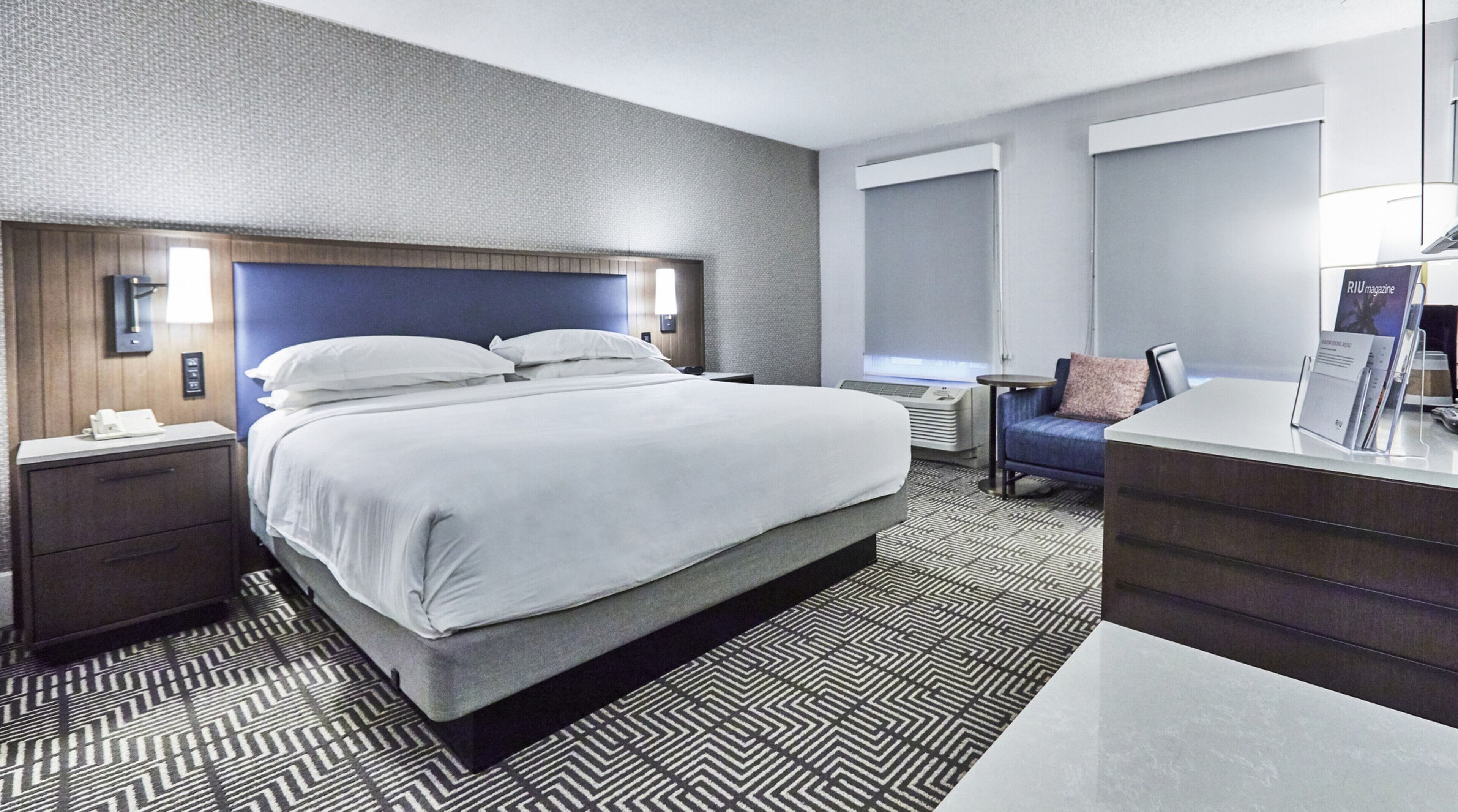 A modern hotel room with a large bed, a desk, a chair, and a small table.